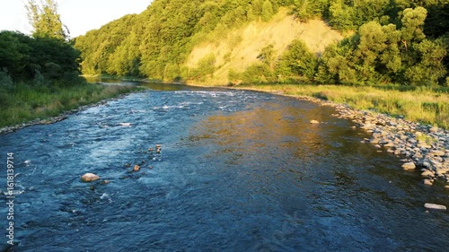 Low-altitude drone view of a calm mountain river flowing gently at golden hour. Warm sunlight illuminates rocky riverbanks and natural landscape, creating a peaceful and cinematic atmosphere. Scenic w