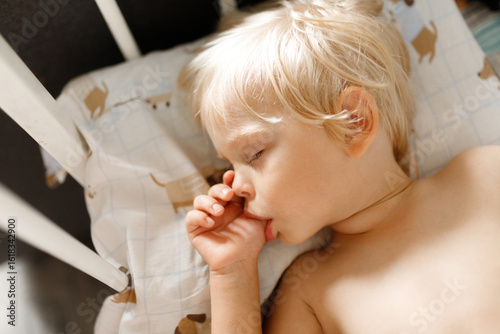 small cute blonde child sleeping in his white crib and sucking his thumb finger. sucking reflex