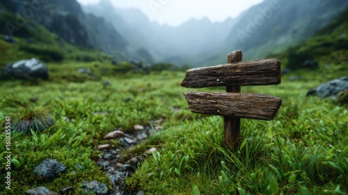 The image depicts a rustic wooden signpost standing amidst lush green grass and rocky terrain in a misty mountain landscape, evoking a sense of tranquility and adventure.