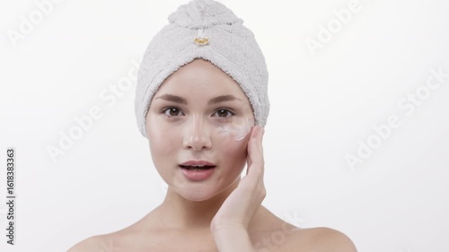 Cute young woman with towel on head applying moisturizing cream to face. Concept: skincare and youth.