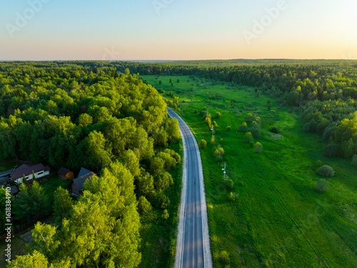 An aerial view shows a winding road cutting through a vast expanse of lush green forest and open fields under a clear sky with a sunset glow