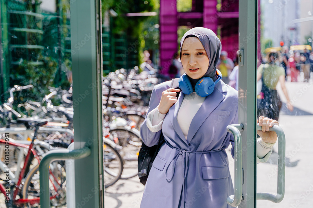 Obraz premium Young Muslim woman exiting building with headphones and backpack in city
