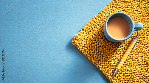 Minimalistic flat lay with a knitted yellow table mat, a pen, and a cup of coffee on a blue background with copy space for text or design.