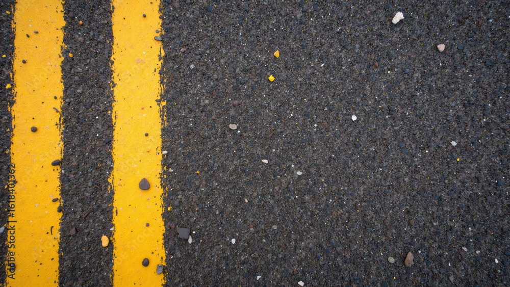 Fototapeta premium Close up view of textured asphalt road featuring two parallel yellow lines, surrounded by small stones and debris, creating striking visual