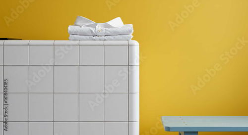 A neat stack of freshly laundered and folded white shirts sits on a white tiled surface against a vibrant yellow background. Minimalist domestic scene.