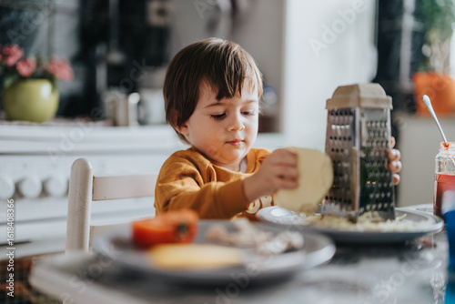 Photos A young child actively grating cheese on a plate while sitting at a kitchen table, creating a warm and homey atmosphere