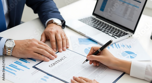 Two business professionals finalizing an agreement by signing a contract document on a desk with financial charts and a laptop.