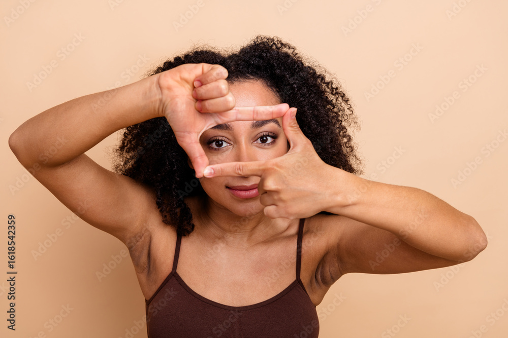 Fototapeta premium Young woman framing her face with fingers against beige background, showing confidence and happiness, trendy casual look