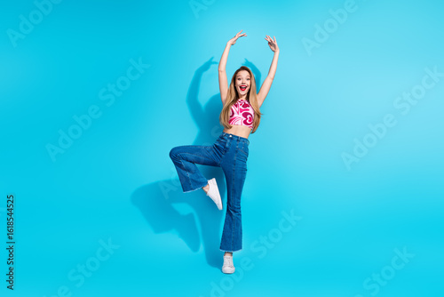 Joyful young woman in trendy outfit posing with arms raised on a vibrant blue background, expressing happiness and energy