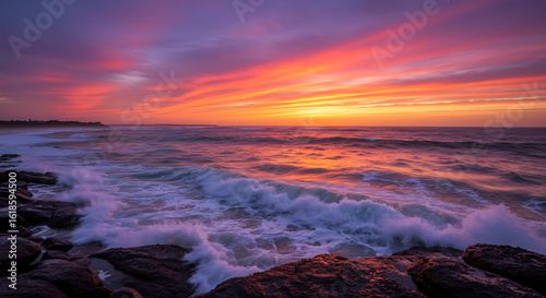 Dramatic coastal sunset with a fiery orange and purple sky reflecting on ocean waves crashing against a rocky shore.