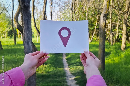 Female hands holding a white sheet of paper with location pin symbol on a pathway in the forest. Lost person travel guidance, discovery, and exploration concepts