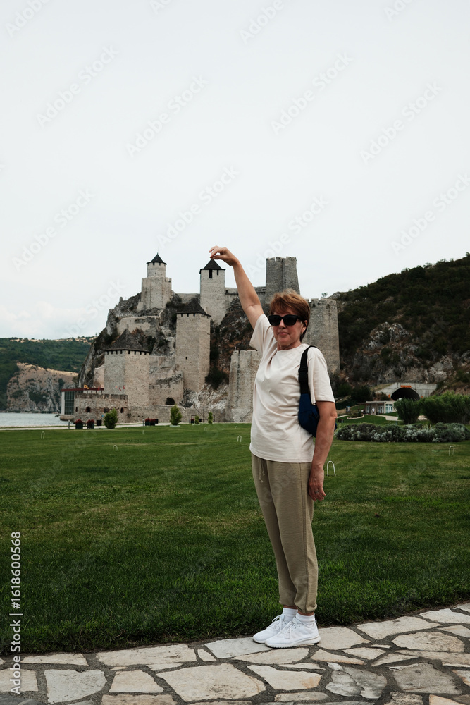 Fototapeta premium Woman posing with raised hand pretending to hold a tower of Golubac Fortress in a playful travel photo. Travel in Serbia concept