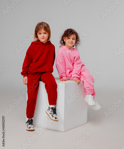 Two young girls are posing together in a studio, showcasing their colorful sweatsuits. One wears a red outfit, while the other is clad in pink, both exuding joy and playfulness.