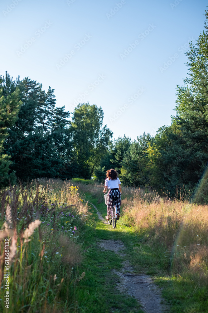 Fototapeta premium Woman riding bicycle along a scenic path through tall grass and wildflowers, surrounded by lush greenery and trees, enjoying a peaceful outdoor experience in nature
