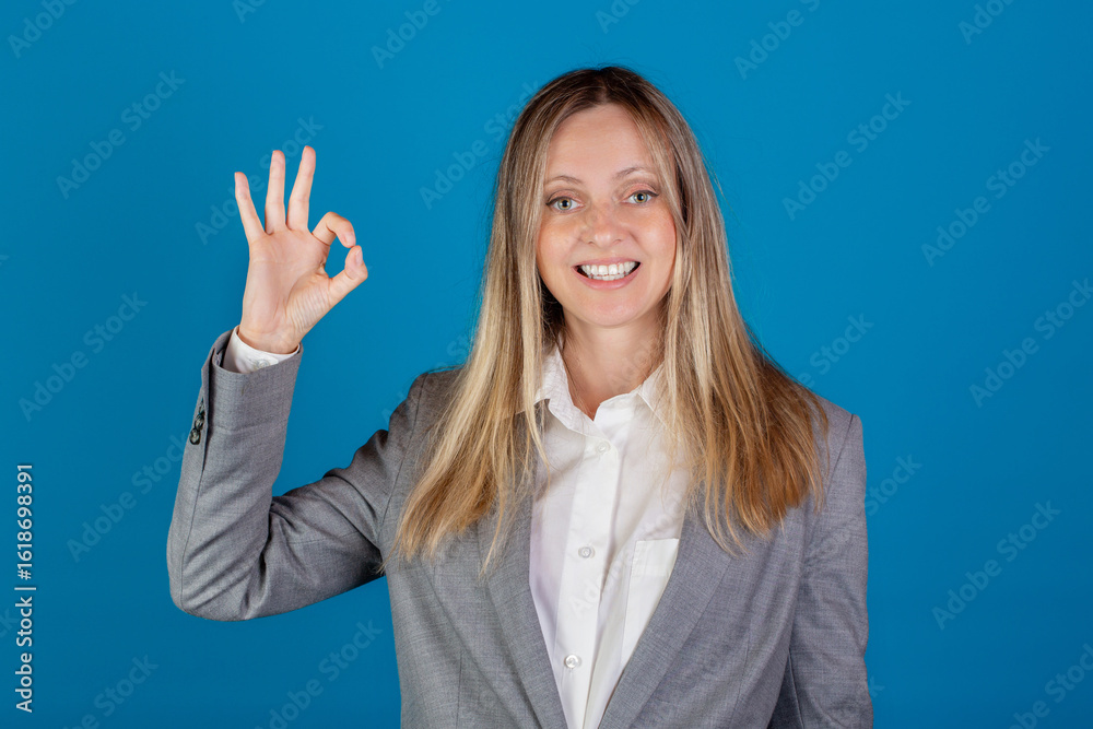 Fototapeta premium Beautiful blonde european caucasian smiling woman in business suit showing OK sign against blue background