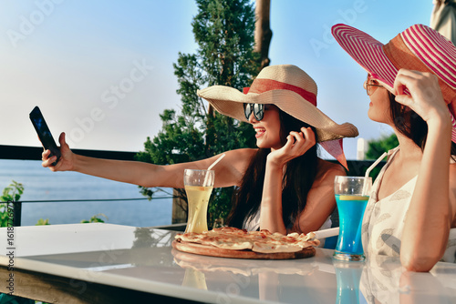 Two friends enjoying cocktails and pizza while taking selfies on a sunny day by the sea.