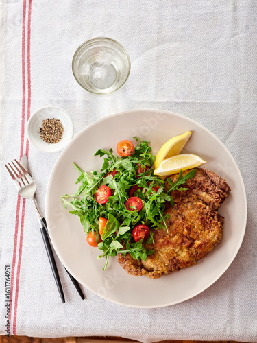 A plate features crispy chicken milanese paired with a vibrant tomato arugula salad and lemon wedges. The meal is beautifully arranged on a textured tablecloth with utensils nearby