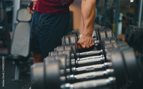 ​A close-up, slightly angled shot of a person's arm and torso reaching for a heavy dumbbell from a rack in a gym.