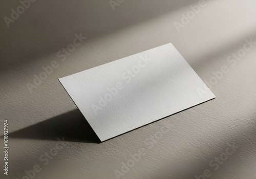 A blank, white business card rests on a textured surface, casting a soft shadow with light filtering from above.