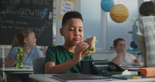 Fotografie African American Primary School Boy Sitting at the Desk, Eating Sandwich from Lunch Box, Talking to Classmate During Break