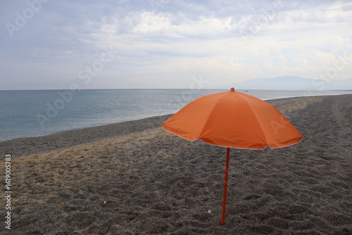 Fototapeta Naklejka Na Ścianę i Meble -  Orange beach umbrella in the rain on a pebble beach by the sea. Calabria, Italy.