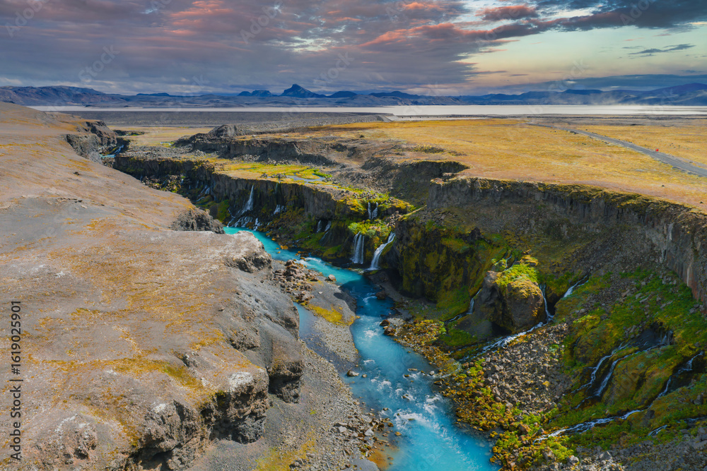Fototapeta premium Sigoldugljufur Canyon in Iceland features a turquoise river, cascading waterfalls, moss covered cliffs, and golden plains under a colorful sunset sky.
