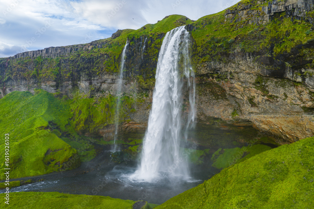 Fototapeta premium Seljalandsfoss waterfall in Iceland flows from a tall moss covered cliff. A walking path near the base and misty spray add to the serene daylight scene.