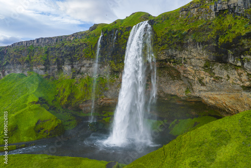 Seljalandsfoss waterfall in Iceland flows from a tall moss covered cliff. A walking path near the base and misty spray add to the serene daylight scene.