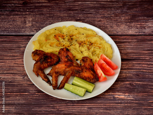 Baked chicken wings with mashed potatoes and pieces of ripe tomatoes and cucumber on a dark wooden table.