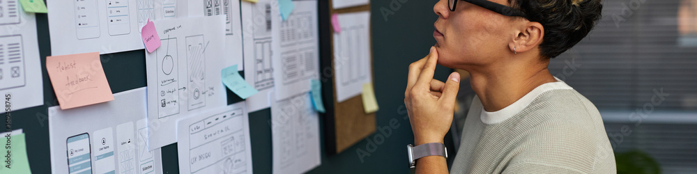 Fototapeta premium Young adult Asian woman analyzing wireframe sketches and sticky notes on wall, standing in modern office, wearing glasses, planning user interface design for digital project