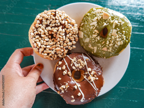 3 Donuts with varios flavour, chocolate, peanut and green tea donuts on the table, green background, top view, served by white plate, held by hand