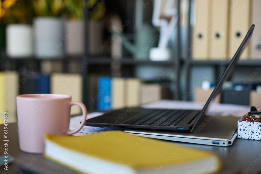 Fototapeta premium Closeup showing laptop, pink mug, and yellow notebook on desk in modern office workspace, blurred shelves with folders and plants in background, no people visible