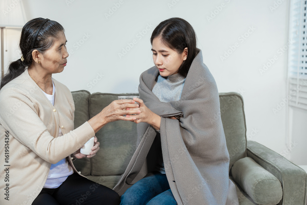 Obraz premium An elderly Asian woman gently checks her sick daughter's forehead, offering care and comfort. The young woman,wrapped in a blanket, holds a glass of water while being looked after in the living room