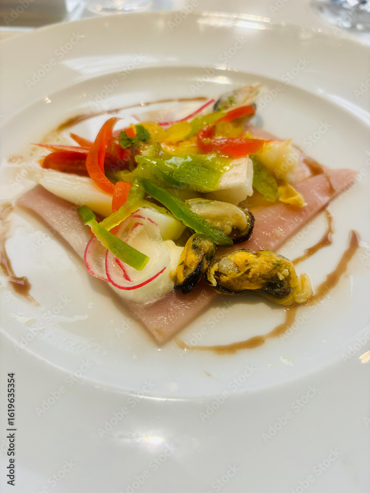 Seafood appetizer plated on a white dish, featuring slices of cooked ham topped with assorted shellfish, squid, and colorful bell peppers. Seafood appetizer plated on a white dish, featuring slices of cooked ham topped with assorted shellfish, squid, and colorful bell peppers.