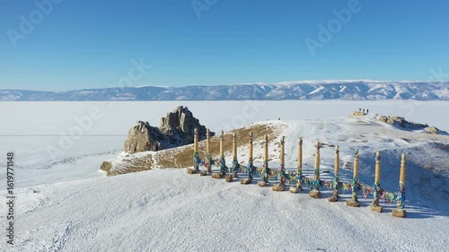 Aerial view of the stark, snow-covered landscape featuring the frozen Lake Baikal, a rocky outcrop, and decorated wooden pillars, Irkutsk Oblast, Russia.