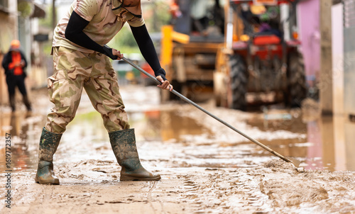 Thai workers cleaning a mud-covered street after a major flood. Wearing safety gear, they work together to restore public roads, showing resilience and community effort in disaster recovery