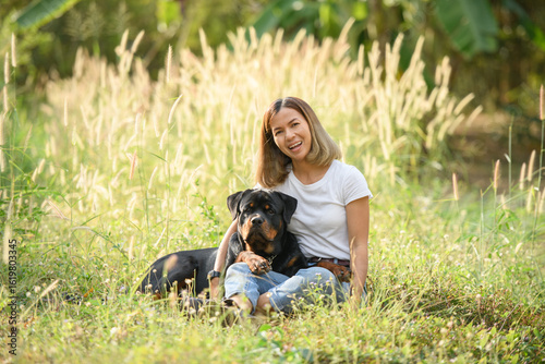 Happy Asian woman sitting in a field with her Rottweiler dog, enjoying nature and sunshine. Concept of pet love, outdoor lifestyle, and peaceful countryside moments.