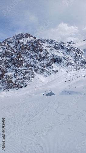 Snow-covered landscape at the Shymbulak ski resort in Kazakhstan, dominated by a rugged mountain range. A semi-circular dome structure, covered in snow, subtly blends into the icy environment