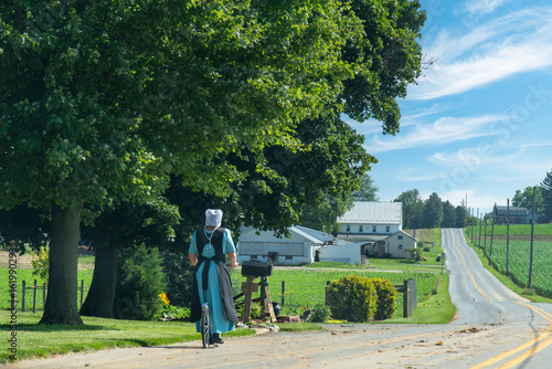 Amish woman on a kickbike or footbike on a rural road surrouned by trees, farms and agricultural fields in the Lancaster area, PA, USA