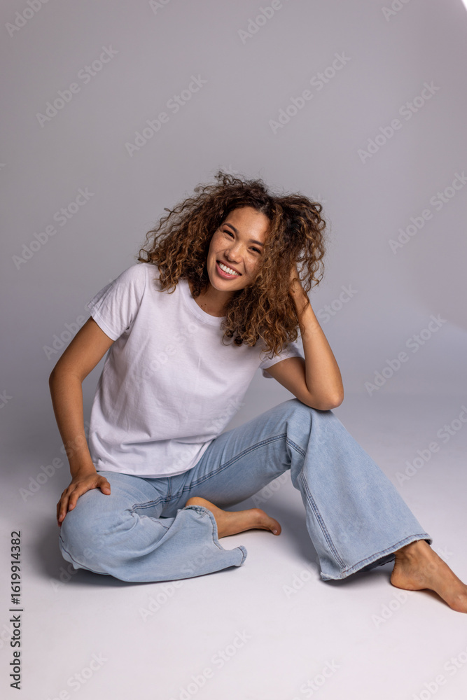 Fototapeta premium Curly Woman Sitting On Floor In White T-Shirt And Blue Jeans