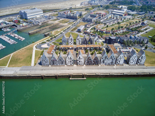 Aerial view of modern, geometric buildings lining the waterfront, contrasting with the bright green water and distant sandy beach, Dunkirk, France.