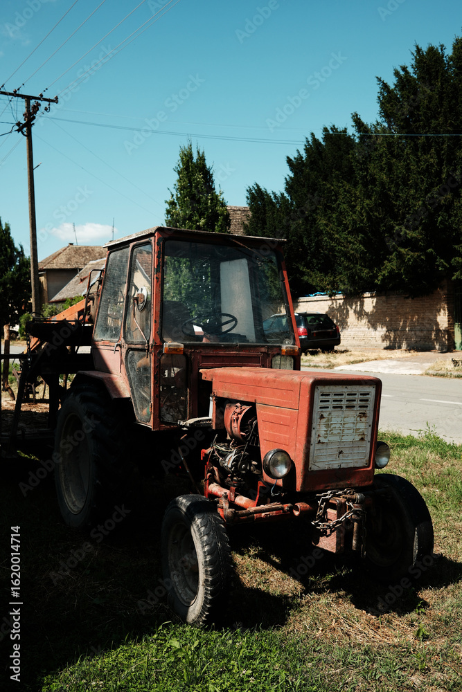 Fototapeta premium Old red tractor with a trailer parked on grass in a residential street in Apatin, Serbia, on a sunny day.