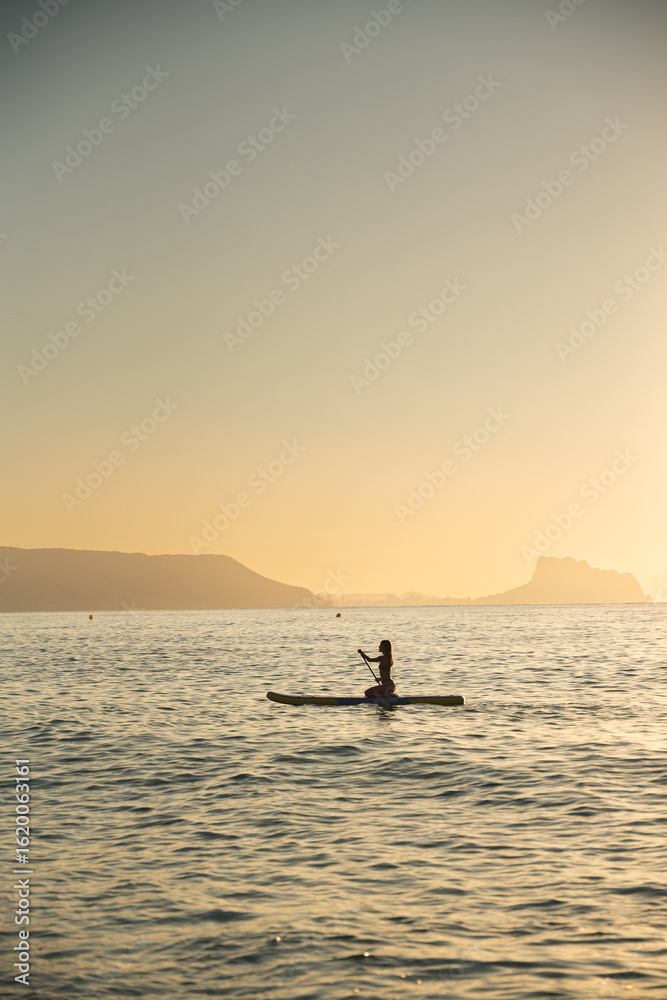 Fototapeta premium Young woman paddleboarding in serene ocean at sunset