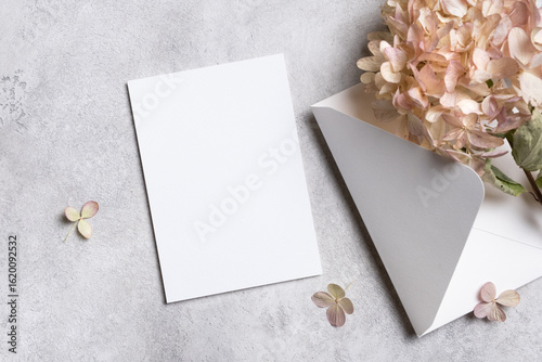 Blank greeting card mock up with white envelope and dried hydrangea flowers on textured gray background, perfect for personalized messages and creative designs