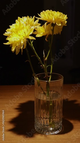 In a dimly lit room, a yellow chrysanthemum rests in a glass on a wooden table, illuminated by the warm glow of a nearby lamp