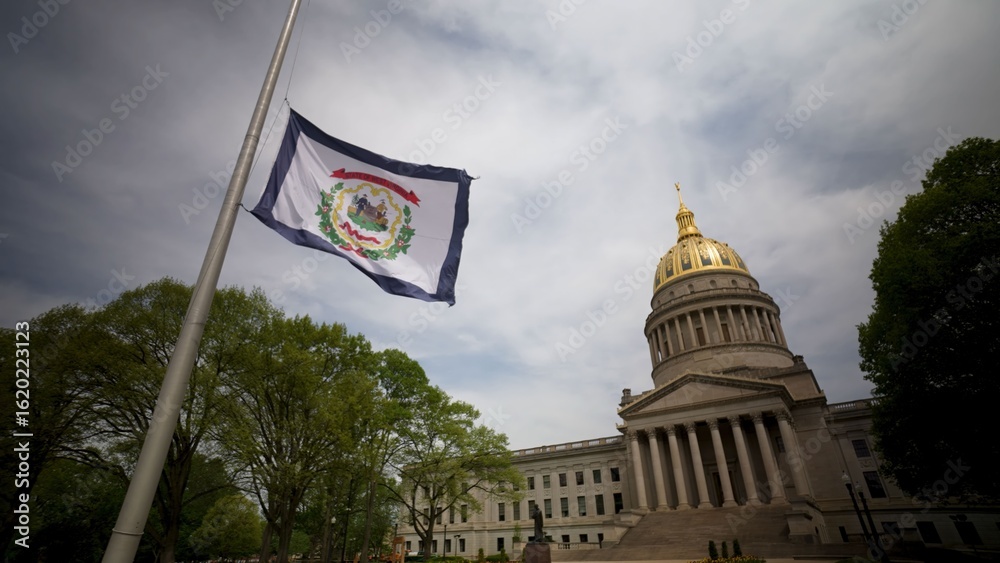 Fototapeta premium The West Virginia State Capitol showcases its impressive dome while the state flag waves gently. Surrounding trees add natural beauty to the historic site in Charleston.