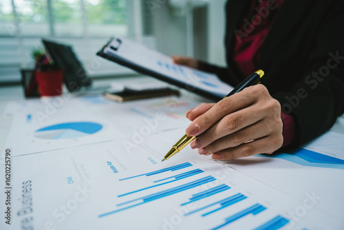 Fotografie A businesswoman analyzes financial documents with charts and graphs at a desk, highlighting data trends with a pen in a bright, professional office setting