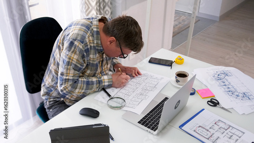 A nerd with glasses hunched over his architectural masterpiece. A man is sitting at a table with a laptop and building drawings