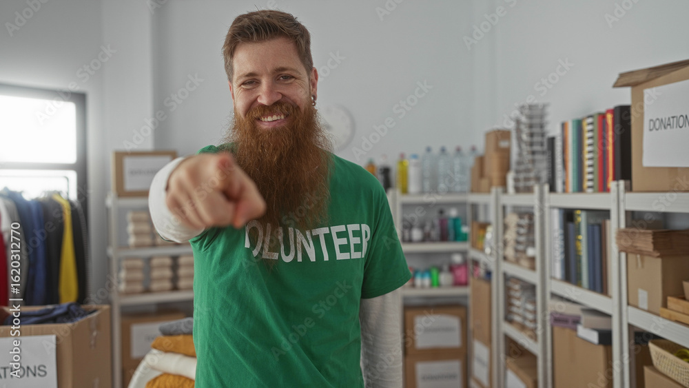 Fototapeta premium Bearded man pointing finger in donation building beside clothes box and shelves; support motivation.