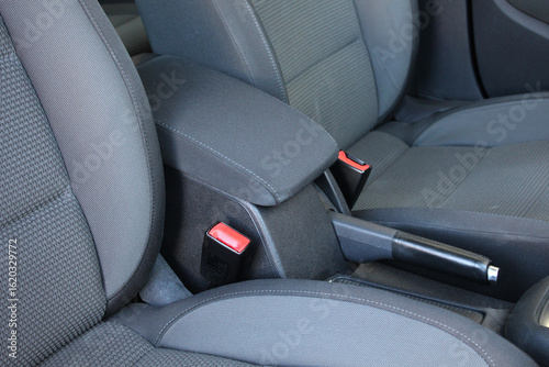 Armrest of a modern car located between the front seats. Detailed view of cars center console: armrest with stitching, seat belt, handbrake with leather handle and seats with gray fabric upholstery.